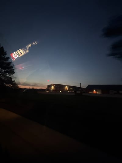 The image depicts an exterior scene at twilight or dawn in Senokos, Bulgaria, captured from within a vehicle. The sky graduates from a dark blue overhead to an orange and pink hue along the horizon. A prominent reflection on the vehicle's window in the upper left quadrant displays a brightly lit interior, featuring a red sign with white lettering visible as "COLA" and what appears to be illuminated shelving. The lower foreground consists of a dark, curved surface consistent with a vehicle's interior. To the far left, the silhouette of a coniferous tree is present. In the midground, across an unlit expanse of ground, two industrial-style buildings are visible. The left building is a multi-story structure with multiple illuminated windows and several bright lights along its roofline; a dark chimney rises behind it. The building to the right is a single-story, darker structure with one external light illuminating a section of its facade. A faint red light source is visible in the sky above the horizon, situated between the reflection and the industrial buildings. No individuals are discernible.
