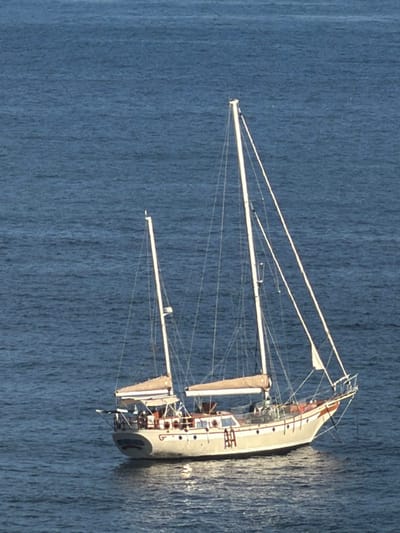 A white sailboat with tan sails drifts on dark blue water. The sails are furled around two spars. The boat appears to be a vintage design with a tall mast. Several people are visible on the deck. The water has a distinct textured appearance.