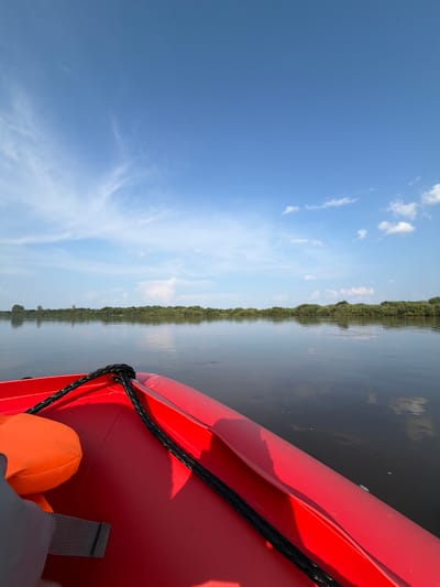 The image shows a first-person perspective from a red kayak on a calm body of water, likely a river, with a dense line of green trees and vegetation forming the far bank. The sky is a clear blue with scattered white clouds. An orange life vest is visible on the left side of the kayak, and a black rope is secured to the front. There are no people visible in the image, and no specific details pinpoint the location to the city of Kalino, Russia. The scene depicts a peaceful, natural environment during daylight hours.