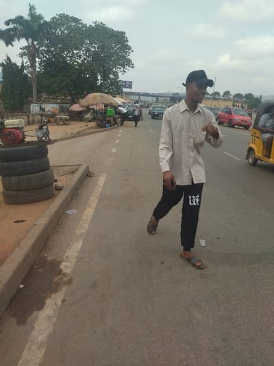 The image shows a man walking down a street in Maraba, Nigeria. He is wearing a light-colored long-sleeved shirt, black pants, a black hat, and sandals. He is looking to the left. The street is paved and has a white painted line down the center. There are several vehicles parked on the side of the road. There is a pile of tires on the right side of the image. The man is walking toward the left. There is a small stand with goods on the right side of the image. There are several other people walking in the distance.  The sky is overcast and there are trees on the side of the road. The scene is in the middle of the day, and the sun is shining.  The man is the subject of the image.