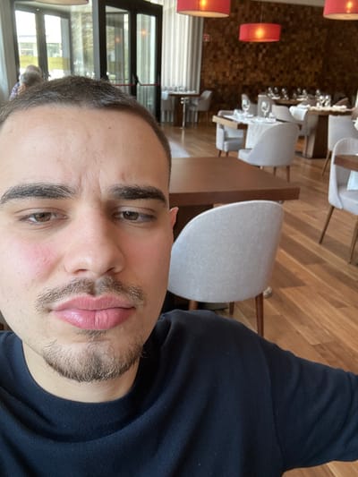 A male subject is prominently featured in the foreground, taking a self-portrait within a modern restaurant environment. He has short, dark hair with some visible grey, dark eyebrows, a mustache, and a chin beard. His complexion shows slight redness on the cheeks, and his lips appear full. He is wearing a dark blue crew-neck long-sleeved garment and looks directly into the camera with a neutral expression.
The background depicts the interior of a sophisticated dining area, identified as the Restaurante Aquae Flaviae within the Hotel Aquae Flaviae in Chaves, Portugal. The room features a light-colored wooden floor with wide planks. The left side of the room includes large glass doors or windows, through which natural light enters and blurred figures are partially visible in the distance. The right wall is accented with a textured wood paneling, composed of small, irregular wooden blocks. Three spherical or dome-shaped red pendant lights are suspended from the ceiling, casting a warm glow. Numerous dining tables are arranged throughout the space, each with a dark brown top and light grey upholstered chairs with wooden legs. Many tables are formally set with white placemats, multiple glasses, and white plates. The restaurant appears largely unoccupied at the time of the photograph.