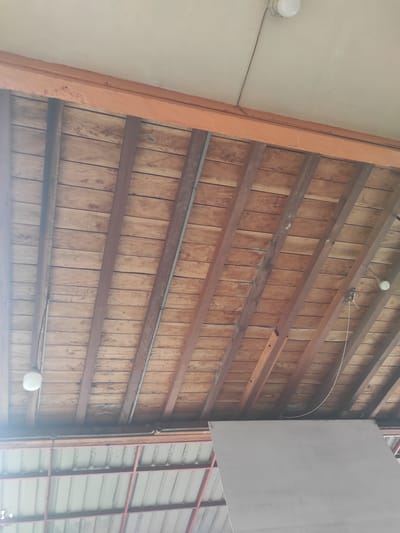 The image displays an upward view of an architectural structure, likely a ceiling or high wall, set in Manetunong, Indonesia. The dominant feature is a rustic wooden surface composed of numerous vertical light-brown wooden planks. These planks are horizontally traversed and supported by several darker, unpainted or stained wooden beams.
Running along some of these wooden beams are grey metal conduits or pipes, suggesting electrical or utility lines. Two spherical white light fixtures are visible, one in the upper-middle section and another towards the lower right, both emitting a soft glow and attached to the wooden structure. A thin white wire arches across the upper right section of the wooden ceiling.
To the left, a thick, vertical beam painted in a vibrant orange hue acts as a structural boundary, separating the wooden ceiling from a plain, light-colored wall surface. On the far right, a large, rectangular white panel hangs vertically, partially obscuring a background structure. Beyond this panel, a framework of reddish-brown metal beams supports what appears to be a section of corrugated metal siding or another wall element.
The scene is brightly lit, indicative of daytime. There are no people, explicit activities, or visible text within the frame. The overall impression is one of utilitarian construction, emphasizing natural wood and functional elements.