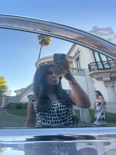 The image captures a mirror selfie taken from within a vehicle, reflected on a slightly smudged window or mirror surface. In the foreground, a woman with long dark hair, dark nail polish, and a black and white patterned top or dress, holds a light-colored smartphone with a circular grip in her right hand to capture the reflection. She wears multiple bracelets, including a pink string and beaded styles, on her left wrist, and a gold-colored necklace with an elongated pendant. Her lips are pursed.
The background, visible through the reflection, features a grand, ornate building situated in Lordelo do Ouro e Massarelos, Portugal. This multi-story structure is light-colored, displaying Neoclassical or Romantic architectural characteristics, including prominent columns, decorative cornices, and a sweeping, curved exterior stone staircase with dark railings on its left side leading to an upper-level entrance. A dark wrought-iron balcony adorns an upper window on the right side of the building. To the left, a tall palm tree stands against a clear blue sky, with additional green foliage visible further left. The ground in front of the building is paved with cobblestones. Two other women are visible in the background: one on the right wears a long white dress with a distinct black pattern and carries a dark clutch bag; the other, partially obscured behind the first and near the staircase, wears a light-colored long dress. The scene is illuminated by bright daylight.