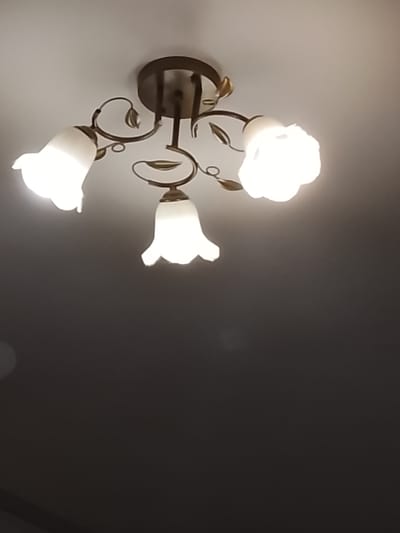 This media file features a close-up, angled shot of a ceiling light fixture. The fixture is made of dark metal with decorative scrollwork and leaf-like accents. It has three lights, each encased in a white, frosted glass shade shaped like a tulip or bell. Two of the lights are on, emitting a bright, warm glow that washes out some of the detail in the surrounding ceiling. The third light is partially visible and appears to be off or dimmer. The ceiling itself is painted a light, neutral color. The lower portion of the image is dark, suggesting it is a dimly lit room or that the photo was taken from a lower vantage point looking upwards. There is no visible text or indication of people or a specific event taking place. The setting appears to be an indoor location, likely a room in a home or building. The lighting suggests it could be evening or a time when artificial light is needed. The overall impression is a functional yet decorative lighting element in a domestic setting.