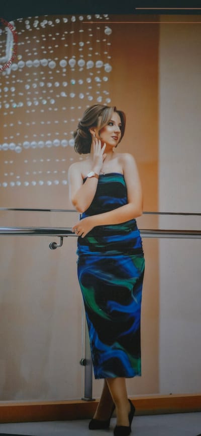 A young woman with her hair styled in an updo stands posing with her hand on her cheek. She is wearing a strapless, floor-length dress with a blue, green, and black swirled pattern. Her left arm is resting on a silver railing. She is wearing a watch on her left wrist and a ring on her left index finger. She is wearing black high-heeled shoes. The background is a light brown wall with a pattern of white circular lights above her. There are no visible indicators of the city of Dankov, Russia.