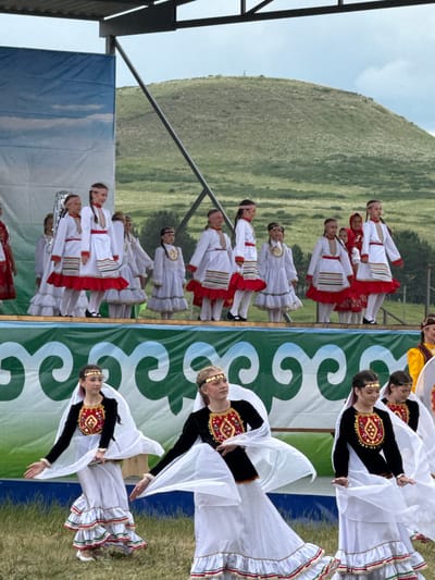The image depicts a cultural performance taking place outdoors in Nurkeevo, Russia. A group of young girls, dressed in traditional folk costumes, are performing on a stage. The costumes consist of white dresses with red and green trim, and some girls wear headbands. The backdrop of the stage features a blue sky with clouds, and behind the stage, a green, grassy hill is visible under a cloudy sky. In the foreground, another group of girls, also in traditional attire, are dancing. Their outfits include black tops with ornate gold and red embroidery, and long, flowing white skirts with ruffled hems. They are also wearing head adornments. The ground is covered in grass, and the overall scene suggests a festive cultural event.