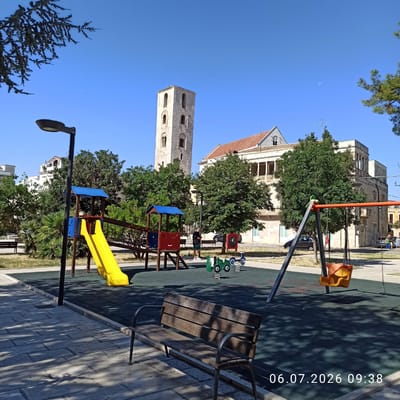 The image depicts a playground area in Ruvo di Puglia, Italy, on July 6, 2026, at 09:38. In the background, a prominent stone bell tower with arched windows stands against a clear blue sky. To the right of the tower is a building with a red-tiled roof and balconies.
The foreground is dominated by playground equipment. A yellow slide descends from a multi-colored play structure. To the right, a swing set with an orange seat is visible. A green spring rider resembling a toy tractor is also present. A wooden bench with metal legs is positioned in the lower part of the frame, casting shadows from nearby trees.
A person in dark clothing and shorts stands near the play structure, looking at their phone. Another person, partially obscured, is visible in the background on the right. Several cars are parked along the street behind the playground. The ground is a dark rubberized surface, bordered by a paved walkway with cobblestone patterns. Lush green trees and some palm trees surround the playground, indicating a public park or square. The lighting suggests it is a bright, sunny morning.
