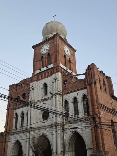 This image shows the facade of a historic church in San Fernando, Chile, under a clear, pale blue sky. The church, constructed primarily of red brick, features a prominent clock tower with two visible clock faces. The tower is topped by a light-colored, dome-like structure with a small cross. The church's architectural style includes arched windows, decorative white plasterwork, and visible signs of damage and disrepair, such as cracks in the plaster and missing sections of the roofline, particularly on the right side of the building. Numerous black electrical wires are strung across the front of the church, obscuring parts of the facade. The lower portion of the church features large arched entrances. The overall impression is of an aging, possibly abandoned or undergoing restoration, religious building in an urban setting.