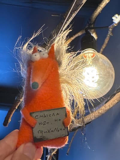 A close-up, slightly blurry, horizontal shot captures a handmade orange felt toy fox in the foreground, held by a person's thumb and index finger. The toy is roughly anthropomorphic, with two large white eyes, a small green nose, and shaggy light brown hair. It holds a small, dark rectangular sign with handwritten text in Cyrillic. The sign reads: "Смысла нет, но прикольно" which translates to "There is no point, but it's funny."
Behind the toy, branches of a tree or shrub are visible, intertwined with electrical wires and a large, glowing Edison-style light bulb. The background is a dark, deep blue, suggesting an indoor setting. The image is lit by the bulb, casting a warm glow on the toy and branches. The overall scene is casual and artistic. There is no specific indication of the location within the city of Kirishi, Russia.
