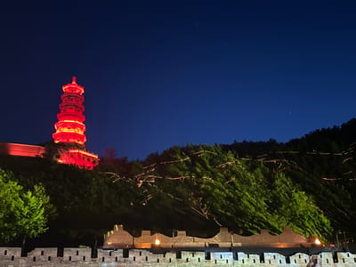 Here is a detailed description of the image:

The image shows a nighttime scene featuring a tall, illuminated pagoda in Miyun District, China. The pagoda, glowing red, stands atop a hill, with a building adjacent to it. A forested hill extends to the right of the pagoda, with strings of lights winding through the trees. In the foreground, a brick wall with crenellations and lit segments runs horizontally across the frame. The sky is a deep blue, suggesting it is nighttime. There are no people visible in the image. The overall scene suggests a tourist attraction or historical site illuminated for nighttime viewing.