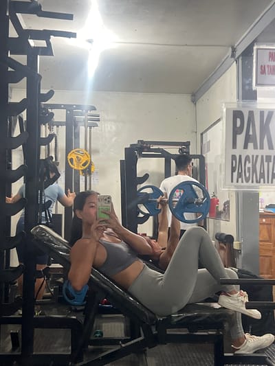 In Cuyo, Philippines, a woman is captured in a mirror selfie at a gym, holding her phone to take a photo. She is wearing a gray sports bra and gray leggings, her hair pulled back. She is positioned on a bench press, with a barbell visible above her. To her right, a man is lying on the bench press, lifting the barbell. In the background, other gym equipment is visible, including weight plates and a squat rack. A sign with text in a foreign language is mounted on the wall. The gym appears to be indoors with fluorescent lighting.