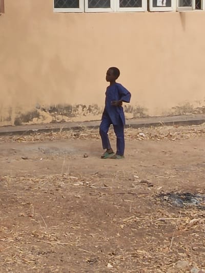 This image, captured in Kanti, Nigeria, shows a young male individual standing outdoors. The boy, with dark skin and short dark hair, wears a dark blue traditional long-sleeved tunic and matching trousers, along with green slide sandals. He stands with his body angled slightly right, facing towards the right side of the frame, his right hand resting on his lower back. In the background, a building with a light orange-tan colored wall is visible, featuring two white-framed windows with diamond-patterned grilles. The lower portion of the wall exhibits discoloration and weathering. The ground is dry, light brown earth, sparsely covered with scattered dried leaves and small debris. A small, dark, irregular object is situated near the boy's left foot. To the right in the foreground, a distinct patch of dark, possibly burnt residue or ash, lies on the ground.