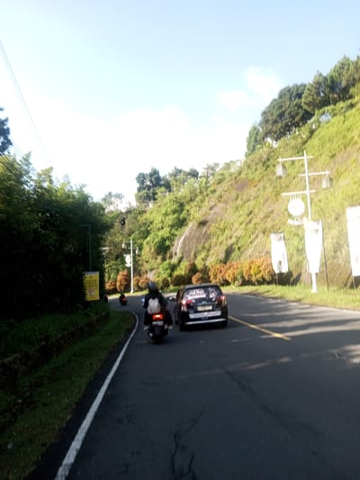 A daytime outdoor shot captures a curved paved road with two motorcycles and a dark-colored car moving in the same direction. The roadside is lush with greenery, including trees and grassy slopes. Utility poles and signage are visible along the roadside. The scene appears to be in a rural or hilly area with clear sky overhead. There is no definitive evidence to confirm the location in Phouviang-Gnai, Laos.