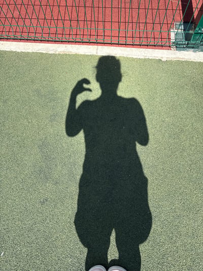 The image shows a shadow of a person on a green, textured outdoor surface, likely a sports court. The shadow depicts the person from the waist down, with one arm bent at the elbow and the hand raised as if to take a photo or make a gesture. The person appears to be wearing shorts. In the background, there is a red surface behind a green metal fence, casting shadows on the red surface. The scene is outdoors under sunlight, indicated by the distinct shadows. The location in the city of Vyazniki, Russia, is not explicitly depicted in the image itself, but is provided as contextual information.