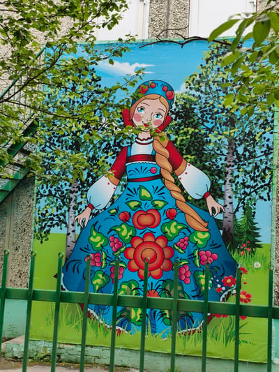 This is a photograph of a mural depicting a woman in traditional Russian dress, possibly a Matryoshka doll, in a stylized natural setting. The mural is situated on the side of a building in the city of Apatity, Russia. The woman has a blue and red head covering adorned with red spheres, blue eyes, rosy cheeks, and a long blonde braid. She wears a red blouse with white puffy sleeves, a blue dress decorated with large red flowers, green leaves, and red berries. The background of the mural features a bright blue sky with white clouds, birch trees with dark bark and green foliage, and a small evergreen tree. The foreground of the mural shows green grass with small red and white flowers. A green metal fence is positioned in front of the mural, partially obscuring the lower portion of the artwork. Lush green foliage from nearby trees hangs down over the top of the mural and the building. The building wall behind the mural appears to be constructed of concrete blocks, with some sections painted white. The mural is an outdoor public artwork.