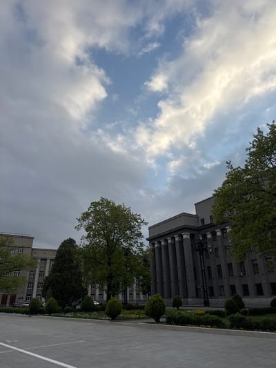 The image captures a view of a building in Akhieli, Georgia, situated under a cloudy sky. The building, constructed with grey materials, features classical architectural elements such as columns. Trees with green foliage are visible on either side of the building, with a paved area stretching out in the foreground. No people or explicit interactions are present. The environmental conditions suggest a possibly overcast day.