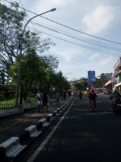 The image shows a street scene during daylight. On the left side of the road is a sidewalk with pedestrians. There is a fence separating the sidewalk from a grassy area with trees. On the right side of the road are motorcycles and cars. In the background, there is a building with a sign that reads "PALACE HOTEL PATAGES". There is also a billboard for "LA Ice". Overhead are power lines and a street light. The overall impression is of a moderately busy street in a city. The city is NOT Phou Khoun, Laos.