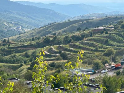 In Uchterek, Uzbekistan, a panoramic view captures the mountainous terrain dotted with residential and industrial structures. In the foreground, leafy branches of deciduous trees obscure parts of several low-rise buildings with dark roofs. Further up the slope, a cluster of larger buildings, including a multi-story structure, is visible nestled among the trees. The midground reveals terraced hillsides covered in greenery, with some areas appearing cultivated. Various industrial elements are present, including a white passenger car parked on a road, a tanker truck with an orange tank, and several large, light-colored spherical tanks. A scattering of trees and shrubs cover the undulating landscape. In the background, multiple layers of hazy blue mountains recede into the distance, indicating a vast and expansive geographical setting.