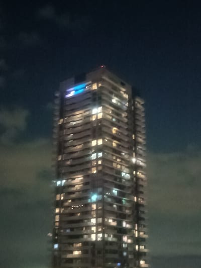 The image shows a close-up, slightly blurred view of a modern, tall, high-rise building at night. The building is illuminated by numerous windows emitting warm yellow light, and there is a prominent section near the top with bright blue lights. The sky is dark with some visible clouds. The building's architectural style suggests it could be a residential condominium. Without further context or identifying markers, it is not possible to definitively determine the specific location within Mississauga, Canada.