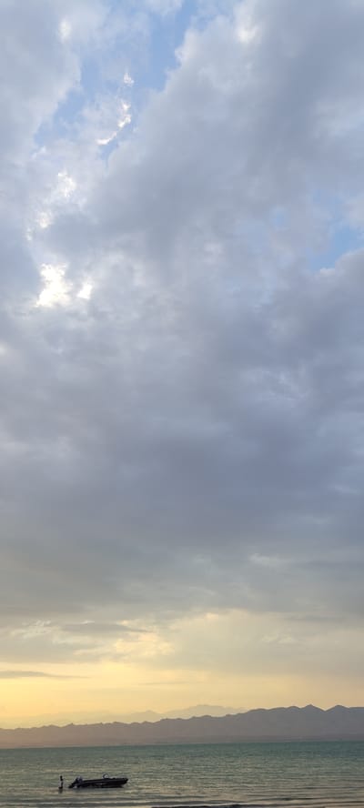 In Chigdalik, Tajikistan, the scene captures a tranquil body of water, likely a lake or sea, with a distant mountain range forming the horizon. The sky is predominantly filled with a layer of soft, gray clouds, with patches of blue sky peeking through and the sun casting a golden glow near the horizon. A small, dark motorboat is visible on the water, with a person standing near it. The water appears calm, with gentle ripples reflecting the muted light. The overall atmosphere is serene and peaceful, suggesting an early morning or late evening setting.