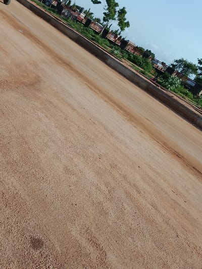This image depicts an outdoor scene in Gbaza, Nigeria. The primary subject is a dirt road or pathway, with a reddish-brown hue. The road occupies most of the frame, extending diagonally from the bottom left to the top right. The surface of the road appears somewhat uneven, with visible textures and possibly tire tracks or other markings.

Along the upper edge of the road, there's a curb or border, transitioning to a grassy area with some vegetation. Beyond this, a fence with red railings encloses a property. Visible within this enclosed area are several structures that resemble market stalls or open-air shops, some with corrugated metal roofs. There are also some trees and foliage. In the distance, a clear blue sky is visible, suggesting it is daytime. The lighting indicates a sunny day, with no clouds immediately apparent.

There are no people clearly visible in the image. The focus is on the road and the area adjacent to it. The overall impression is of a rural or semi-urban environment in Nigeria, likely during daylight hours. The lack of harsh shadows might suggest it is not the middle of the day, or the sun is at an angle that minimizes deep shadows. No text is discernible in the image.
