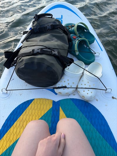 The image shows the surface of a paddleboard on water, likely a lake or river, though the location in Selychka, Russia, is not verifiable from the image alone.

On the paddleboard, there is a dark gray and black dry bag secured with a black bungee cord. Two pairs of Crocs-style shoes are also present: one pair is white, and the other is teal. The right side of the image includes a small portion of a person's legs and hand resting on the board.

The paddleboard is white with blue and yellow striped detailing. Water surrounds the board, suggesting it is in use.