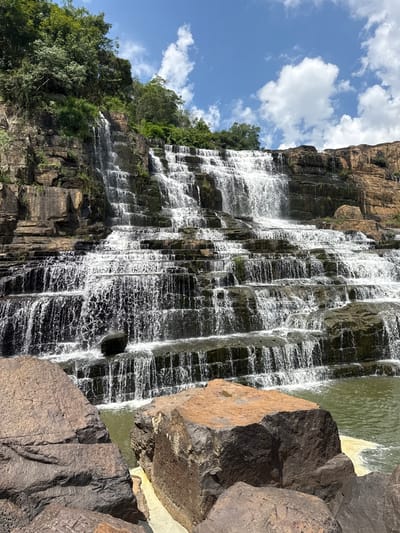 This is a wide-angle, eye-level shot of the Datanla Waterfall, a tiered waterfall in the city of Tân Phú, Vietnam. The waterfall cascades over multiple rock ledges, creating a frothy white curtain of water. Lush green trees and vegetation line the top and sides of the rocky cliffs. The sky is blue with scattered white clouds. In the foreground, large, textured rocks are in sharp focus, with the water of the pool at the base of the waterfall visible behind them. The water appears greenish-brown. There are no people or animals visible in the image. The overall impression is one of natural beauty and serene grandeur.