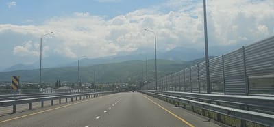 The image captures a view from a highway in Қырғауылды, Kazakhstan, with a mountain range visible in the distance. The highway has multiple lanes and is bordered by guardrails. On the right side of the highway, there is a tall sound barrier made of metal panels and transparent sections. Streetlights are spaced along the highway. In the middle ground, there's a settlement with houses and trees. The sky is partly cloudy. The overall scene depicts a section of road infrastructure in a semi-urban or suburban area adjacent to natural landscapes.