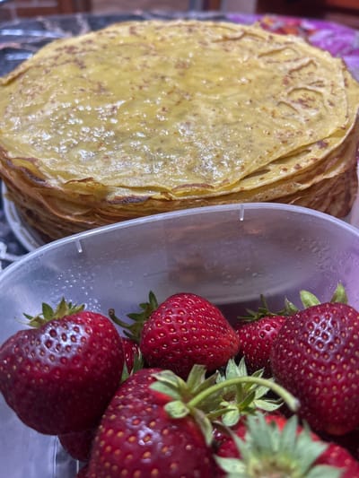 A stack of golden crepes sits behind a clear plastic container filled with fresh strawberries. The crepes are thin and layered, with some browning visible on the edges. The strawberries are ripe and red, with green stems and leaves. The container is partially filled with what appears to be condensation. The background is blurred, but hints of a dark, possibly marble countertop and a brick wall can be discerned. This scene is not specific to Gogino, Russia, or any other city.