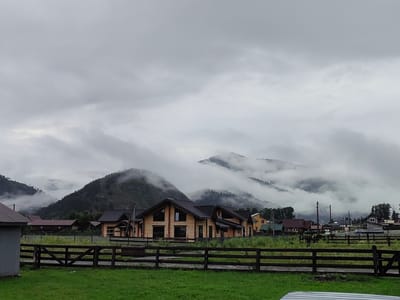 The image depicts a rural landscape in Anos, Russia, under a cloudy, overcast sky. In the foreground, a lush green grassy area is bordered by a dark wooden fence. Beyond the fence, several modern wooden houses with dark roofs are visible. The houses are clustered together, suggesting a small village or a collection of residences. In the background, rolling hills or mountains are covered in dense green trees, and a thick layer of fog or low-lying clouds partially obscures their peaks, creating a dramatic and atmospheric effect. The overall scene conveys a sense of quiet, natural beauty, possibly in a mountainous region.