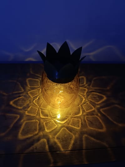 A pineapple-shaped lantern sits on a wooden surface, casting a patterned light on the table and wall behind it. The light is golden, and the room is dark, creating a dramatic contrast. The pineapple's "leaves" are silhouetted against the dark blue background. The origin or location of this image in Cherkovitsa, Bulgaria, is unknown.