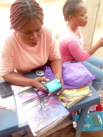 The image captures two women seated, likely outdoors or in a semi-open setting, during daytime. The foreground woman, with reddish-brown braided hair and wearing a light peach-colored short-sleeved top, is centrally focused. She has dark skin and is looking downwards intently at a small, light blue electronic device she holds in both hands. The device has a white emblem or button on its top surface, possibly displaying "WD" or similar letters. Her left wrist has a thin gold-colored bracelet, and her fingernails are manicured with a light polish.
In front of her, on a blue plastic table or stool, are several objects. A dark-colored mobile phone lies on the left side. To its right, a prominent notebook features a serene cover design: a silhouette of a bare tree against a vibrant purple and orange twilight or dawn sky. The words "CAMPUS NOTEBOOK" are clearly visible at the bottom of this cover, along with a partial word "PCTURE" (likely "PICTURE"). Below this, another notebook with a yellowish cover is visible. A crumpled purple plastic bag sits on the table to the right of the notebooks.
Behind and to the right of the foreground woman, the second woman is visible in profile. She has dark hair pulled back, is wearing a pink long-sleeved top and blue jeans, and a small gold hoop earring in her visible ear. She appears to be looking towards the right side of the frame, away from the camera, with a neutral expression.
Below the table, partially visible on the right, is another blue object, possibly a child's learning toy or a small register, characterized by numerous small, colorful circular buttons or pegs. The number "4000" and some other faint text are visible on this object. The background is somewhat blurred, showing what appears to be a light-colored wall or structure. The lighting suggests a bright day.