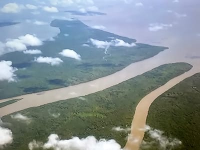 This is an aerial photograph of a river delta or estuary. The dominant subjects are a wide, muddy river and the surrounding land, which is covered in dense green vegetation. The river curves through the landscape, with one section appearing to split and surround a long, narrow peninsula of land. There are scattered white clouds in the sky, suggesting it is daytime and possibly a partly cloudy day. The scene is natural and undeveloped, likely a remote or wild area. There are no people, objects, or text visible in the image. The colors are muted, with the earthy tones of the river contrasting with the vibrant green of the land and the pale blue of the sky. The image has a slightly grainy texture. While the provided location context is Valladolid, Spain, this landscape does not strongly resemble the typical geography of that region.