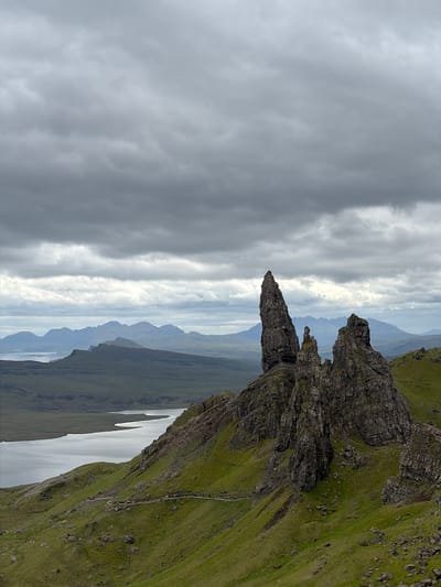 This image captures a wide-angle view of a dramatic, rugged landscape under a heavily overcast sky, identified as the Old Man of Storr on the Isle of Skye, near Portree, United Kingdom.
The upper approximately one-third of the frame is dominated by a dense, unbroken layer of grey stratocumulus clouds, exhibiting subtle variations in tone but no visible patches of blue sky or direct sunlight.
In the mid-ground, slightly to the right of the central vertical axis, stand prominent, dark grey rock formations. The most notable is a tall, slender, spire-like pinnacle, known as the Old Man of Storr, characterized by its irregular, jagged profile and a slight lean. To its right and slightly behind it are several broader, multi-faceted rock stacks and outcrops, appearing as part of a larger, eroded cliff face. These rock structures display visible layering, fractures, and dark, weathered surfaces, with sparse patches of green vegetation clinging to their crevices.
The immediate foreground and mid-ground, particularly to the left and extending across the bottom of the frame, consist of steep, undulating hillsides covered in vibrant green grass. Scattered dark rocks and boulders are visible on these slopes. A winding, light-colored dirt path or trail traverses the green hillside in the lower-left and central mid-ground, snaking its way towards the rock formations. Numerous small, dark human figures are discernible walking along this path, indicating it is a popular hiking route.
To the left of the frame, occupying a significant portion of the mid-ground, is a calm, expansive body of water, likely a loch or sea inlet. Its surface reflects the grey sky, appearing a muted silver-grey. The irregular shoreline follows the contours of the land.
Beyond the loch and the immediate hills, in the distant background, a series of hazy, bluish-grey mountain ranges stretch across the horizon. These distant peaks present a softer, less defined profile compared to the foreground rocks, consistent with atmospheric perspective.
The overall lighting is diffuse and soft, with no harsh shadows, indicating an absence of direct sunlight. The color palette is dominated by various shades of grey (sky, water, rocks, distant mountains) and rich greens (grass and vegetation).