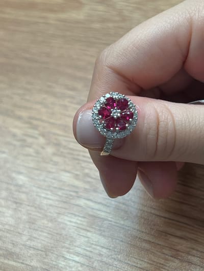A close-up, top-down shot shows a hand, with neatly manicured nails, holding a gold ring with a cluster of gemstones. The ring features a central arrangement of red gemstones, possibly rubies, in a floral or starburst pattern, surrounded by a halo of smaller, clear stones, likely diamonds. The band of the ring is also adorned with small, clear stones. The background is a light-colored wooden surface with a distinct grain pattern, suggesting a table or desk. The lighting is soft and even, indicating an indoor setting. No text is visible in the image. The overall impression is one of elegance and detail, focusing on the intricate design of the jewelry. The cropped images provide clearer views of the textures of the skin, the gemstones, and the wood grain.