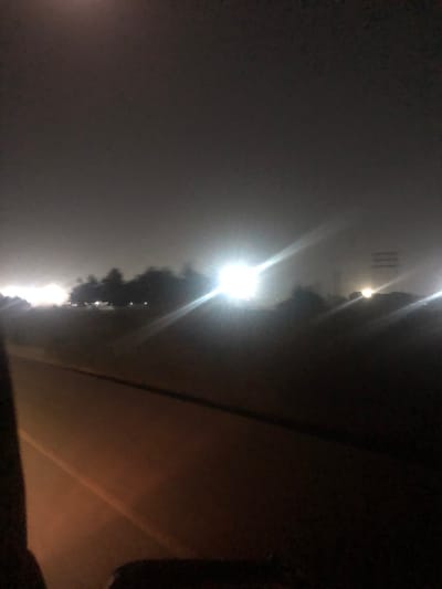 Night scene under a bridge. A bright streetlight illuminates the scene through dense fog, creating a strong glare. Below the bridge, a blurry line of lights is visible, possibly indicating a road or urban area shrouded in mist. The overall ambiance is dark, with limited visibility due to the thick fog.