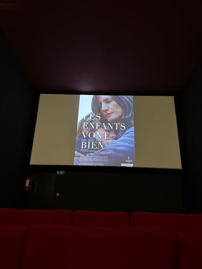 A movie theater displays the film "Les enfants vont bien." The screen shows the movie poster featuring a woman and child. Red seats line the right side of the room. An exit sign is visible near the screen's edge. The room is dark, with the screen's light illuminating the scene.