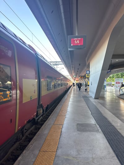 The image shows a train station in Afragola, Italy. The train is red and tan, and is stopped at the platform. The train's coach is 5/6. People are waiting on the platform near the escalators.