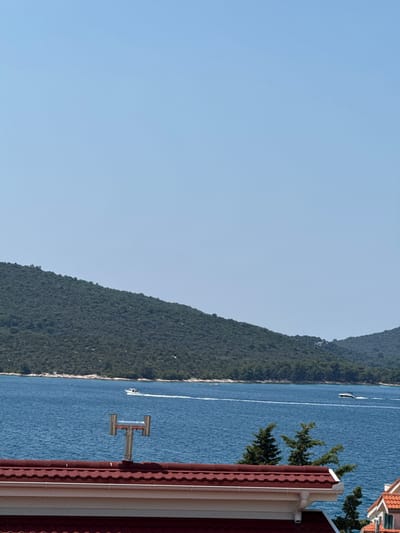 The image shows a view of the Adriatic Sea from a rooftop in the city of Šibenik, Croatia. In the foreground, a section of a red-tiled roof with a white gutter and a metallic chimney is visible. The sea is a deep blue, with two white boats traversing its surface, leaving white wakes. In the background, a densely forested hillside slopes down towards the water, forming the coastline. The sky is clear and bright blue, indicating a sunny day.