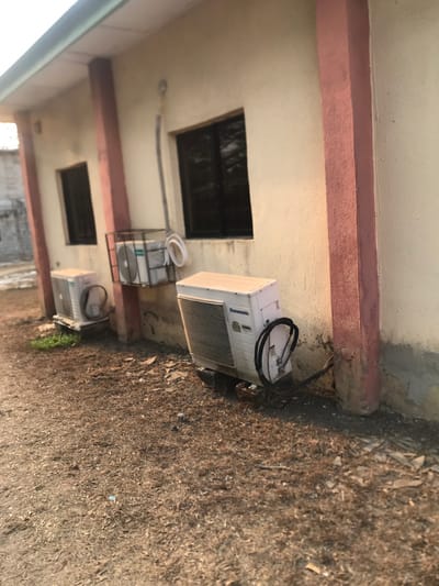 The image displays an exterior view of a building wall and the dry ground beside it, set in Umudike, Nigeria. The building features a light-colored, possibly cream or beige, wall with a prominent reddish-brown horizontal beam or trim running across it. Two dark, rectangular openings, resembling windows or vents, are visible in the wall.
Lining the base of the wall, on the ground, are three outdoor air conditioning units. The most prominent unit, positioned towards the top right of the frame, is white and bears the brand name "Panasonic" clearly visible on its side. Black hoses or cables are coiled on top of this unit. Further down the wall, another air conditioning unit is partially enclosed within a black metal cage or frame, with white tubing or coils nearby. A third similar white unit is visible further along the wall towards the bottom of the frame. A grey electrical conduit or pipe runs along the wall, connected to one of the units.
The ground adjacent to the building is dry and covered in sparse, light-brown vegetation, appearing like dry grass or dirt, interspersed with small rocks and some scattered debris. The lighting suggests it is daytime, with clear weather conditions. There are no people visible, and no specific activity is taking place in the scene.