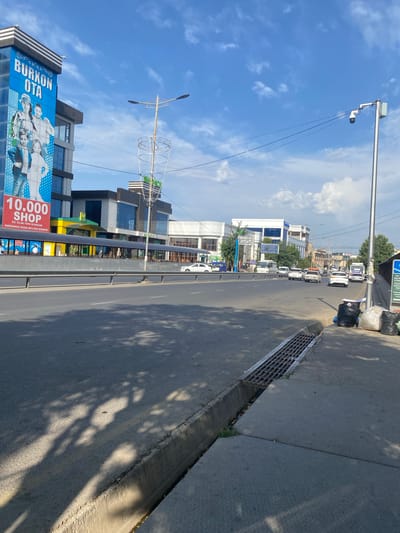 This is a daytime, eye-level, outdoor, street-level photograph taken in Parkent, Uzbekistan. The image is oriented vertically, showing a wide road with several cars driving on it, and a sidewalk on the right. The sky is bright blue with some scattered white clouds, suggesting fair weather. Sunlight casts long shadows of trees across the road and sidewalk.
On the left side of the road, there are several modern buildings with glass facades and some signage. One prominent billboard displays an advertisement for "BURKON OTA" and "10.000 SHOP". The text on the billboard is in a local language. Further down the road, more buildings and trees are visible. A traffic light and a street lamp with a camera are also present.
On the right side of the road, the sidewalk is made of concrete and has a metal grate drainage system running along its edge. A few black and white trash bags are piled up next to the sidewalk. The overall scene depicts a typical urban street environment during daylight hours. There is no discernible activity or event taking place beyond the normal flow of traffic. The mood is neutral and observational.