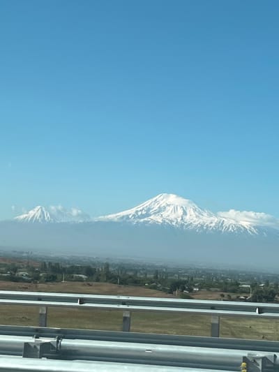 From an elevated perspective, a mountain dominates the scene, possibly Mt. Ararat, with snow-capped peaks partially obscured by clouds. Adjacent to the mountain, a cityscape fades into the horizon, likely Agarak, Armenia. A highway guardrail occupies the foreground, suggesting the image was taken from a moving vehicle on a highway.