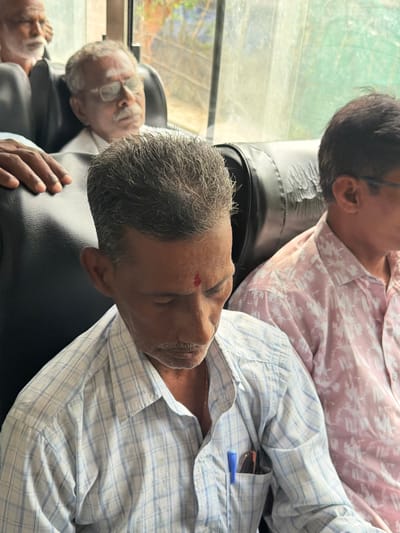 The image shows three men sitting inside a bus in Madirimangalam, India. The man in the foreground is wearing a blue and white checkered shirt and has a red mark on his forehead. He is looking down and has a mustache. To his right, another man is wearing a pink shirt with a floral pattern and glasses. He is looking to the side. In the background, a third man with glasses and a white shirt is visible. The bus appears to have black leather seats and the window is covered with a light-colored curtain.