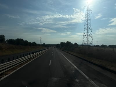 The image depicts a highway in Follonica, Italy, under a bright, partly cloudy sky. The sun is shining intensely, creating a lens flare effect in the upper right portion of the frame and casting long shadows. The highway appears to be a multi-lane road with clear lane markings, suggesting it's a route for vehicular traffic. Guardrails are visible along the left side of the road, separating it from a grassy embankment.

Dominating the right side of the scene is a tall, intricate electricity pylon, its metallic structure silhouetted against the sky. More power lines stretch across the frame, connecting to other pylons in the distance. To the left and right of the highway, dense vegetation and trees line the landscape, with some appearing as dark silhouettes due to the strong sunlight.

The time of day appears to be late morning or early afternoon, given the sun's position and the intensity of light. The weather seems to be pleasant, with the presence of scattered clouds indicating fair conditions, although the harsh sunlight suggests it could be quite warm. No people are visible in the image, nor are any specific activities taking place beyond the implied use of the highway for travel. The overall mood is one of vastness and open road, with a touch of industrial presence from the power infrastructure. There is no visible text in the image.