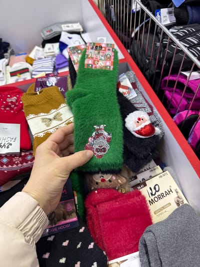 Here is a description of the image:
The image is taken in what appears to be a store in Beloyarsky, Russia. The focus is a shelf filled with various socks. There are Christmas-themed socks in the selection, including designs with Santa Claus, reindeer, and holiday decorations. A person's hand is visible, holding a green, fuzzy sock with a sparkly Christmas ornament. The socks are neatly stacked and displayed in a red-edged shelving unit. There are also packages of socks visible, some with Russian text printed on them, indicating product information. Shelves are present above the socks with clothes.