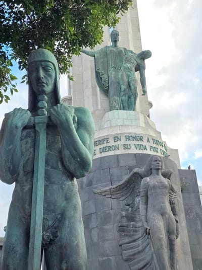 The image shows the Monumento a los Caídos (Monument to the Fallen) in Santa Cruz de Tenerife, Spain. In the foreground, a large, weathered bronze statue of a warrior stands holding a sword. The statue is detailed, with muscular arms and a helmeted head. Behind the warrior, the monument rises, featuring several other statues and inscriptions. A prominent statue on the upper level depicts a figure in robes holding a fallen figure. Below this, a stone inscription reads "JERIFE EN HONOR A TODOS LOS QUE DIERON SU VIDA POR ESPAÑA" (This is a memorial in honor of all those who gave their lives for Spain). At the base of the monument, a winged female statue carved in stone stands with a solemn expression. The sky is cloudy, and lush green foliage from a nearby tree drapes over the top left of the scene. Two birds are perched on the monument.