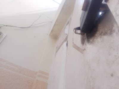 This image depicts a section of a light-colored building exterior or semi-enclosed structure in Chiranchi, Nigeria. A dark, rectangular, possibly electrical or lighting fixture is affixed to an off-white wall in the upper-right quadrant. Directly below this fixture, a light-colored, U-shaped metal bracket is mounted. The wall surface immediately surrounding and beneath the fixture and bracket exhibits significant dark discoloration, consistent with soot, grime, or environmental staining. In the upper-left, multiple thin, black electrical wires are strung horizontally across a clean, light-colored wall. A partial view of a white rectangular electrical box or outlet is visible on the far left edge of the frame. The lower portion of the visible wall surface features a light brown, textured horizontal band, possibly decorative trim or tiling. No individuals are present.