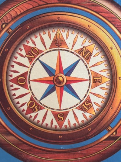 The image displays a close-up, overhead view of a decorative compass, likely a printed illustration or part of a nautical-themed object. The compass features a detailed design with a prominent star-shaped needle in red, blue, and orange. Around the center, cardinal directions are marked with stylized letters on a white background, including "NO" for North, "NE" for Northeast, "E" for East, "SE" for Southeast, "S" for South, "OS" (likely West), and parts of other directional indicators. The compass is surrounded by a textured, wooden-rimmed frame with metallic accents and appears to be set against a blue background. There are no people, animals, or any indication of a specific city or location like Arcahueja, Spain.