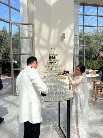 The scene depicts a wedding reception in a brightly illuminated room in La Candelaria, Mexico, featuring a champagne tower. In the center, a multi-tiered pyramid of champagne coupe glasses, largely filled with pale yellow liquid, rests on a round, reflective glass-top table with a black base. A male subject, positioned to the left and viewed from behind, wears a white dinner jacket over a dark shirt and black trousers, actively pouring liquid from a dark bottle into the lower section of the champagne tower. To the right, a female subject, identifiable as a bride, wears a long-sleeved white wedding gown with intricate floral lace patterns and a veil. She pours from a dark bottle with an orange label into an upper tier of the champagne tower.

The room features high, white walls and a light-colored, patterned marble floor. Large, dark-framed, multi-pane windows line the background, offering a view of a sunny outdoor environment with mature olive trees and dense green hedges under a clear blue sky. Sunlight streams through the windows, casting distinct shadows of the window frames and foliage onto the interior wall. A white speaker is mounted on the wall to the right. In the far-right background, a tall, white-linened table with drinks and several wooden bar stools is partially visible, with a person in a dark suit partially seen near it.