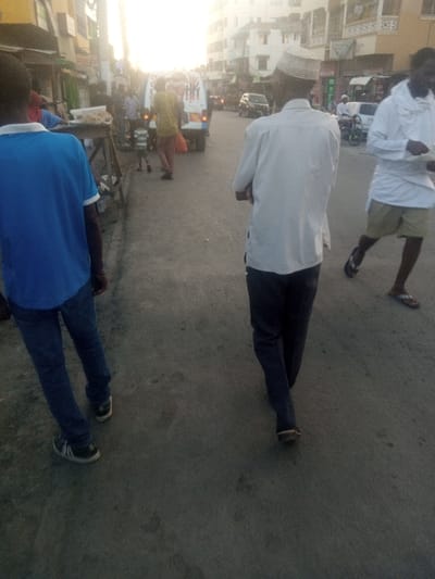 The image depicts an active street scene in Mtopanga ward, Kenya, under low-angle sunlight originating from the background, indicating either early morning or late afternoon. The vantage point is positioned behind several pedestrians on a paved thoroughfare.
In the immediate foreground on the left, a person is visible from the waist down, wearing a blue t-shirt with white trim on the sleeves, dark blue jeans, and black sneakers with white soles, walking away from the viewer. Centrally positioned in the foreground, a man in a white long-sleeved tunic, dark trousers, and a light-colored kufi walks away, with his hands clasped behind his back. On the far right foreground, a partially visible man wears a white long-sleeved top, light-colored shorts, and dark flip-flops, appearing to hold an unspecified item in his hands while walking towards the right.
The midground reveals a bustling street environment. On the left sidewalk, a makeshift wooden stall displays light-colored items, with multiple individuals gathered around it. A person in a yellow shirt and reddish-brown trousers is seen walking past the stall, near a white van with visible rear graphics. A small child is also walking in the vicinity of the stall. Further down the road, a dark-colored car is present, and a motorcycle is visible on the right. Multi-story buildings, consistent with commercial or residential structures, line both sides of the street, featuring windows and various storefronts. The background is significantly overexposed by direct sunlight, which limits detail visibility of distant elements. The road surface is dark, appears paved, and exhibits signs of wear.